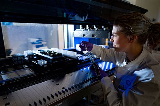 Een laboratoriummedewerker in een witte jas en paarse handschoenen werkt met een pipet bij een high-tech laboratoriumapparaat