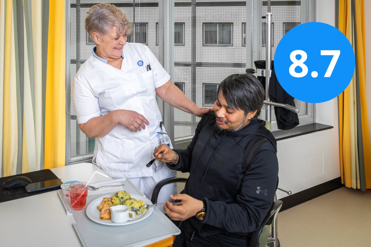 Een zorgverlener met een patiënt met een bord eten. Het cijfer 8.7, waarmee het UMC Utrecht wordt beoordeeld in de Patiëntervaringsmonitor, staat in de rechter bovenhoek.