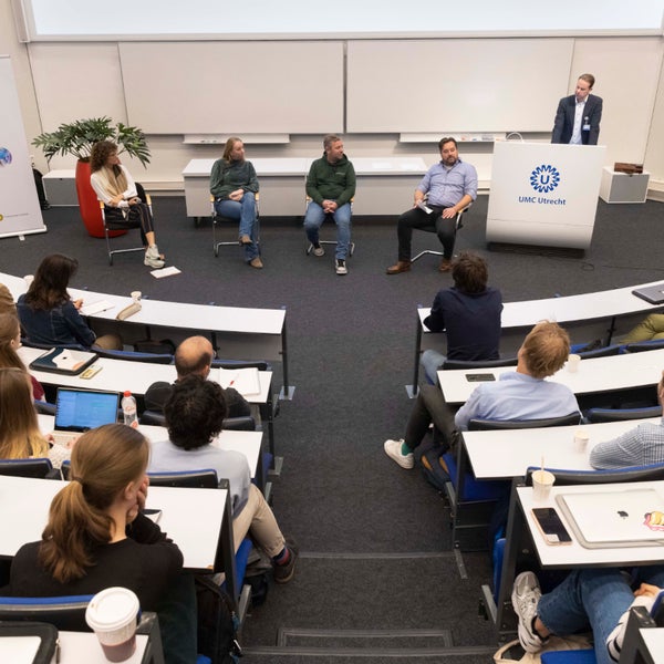 Studenten zitten in een college zaal en kijken naar een groepje mensen op stoelen die in gesprek zijn.