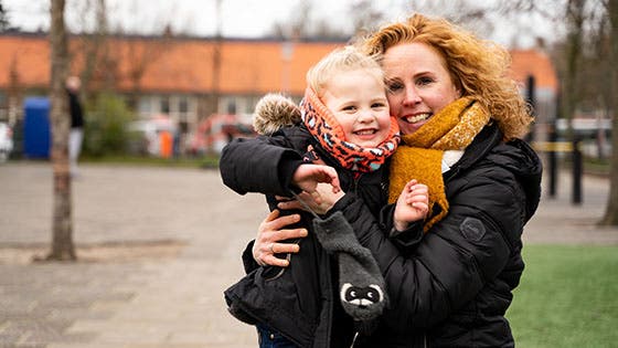 Een moeder houdt haar lachende jonge kind in haar armen buiten op een koude dag, beiden warm aangekleed met sjaals en jassen