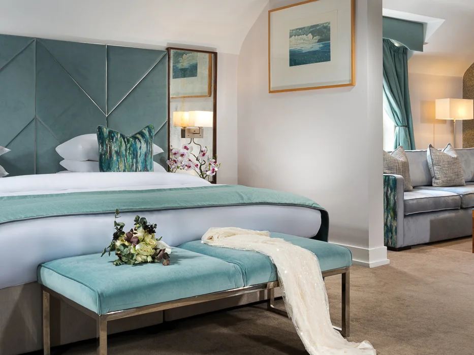 Luxury hotel bedroom with a teal velvet headboard, crisp white linen, and an adjacent seating area