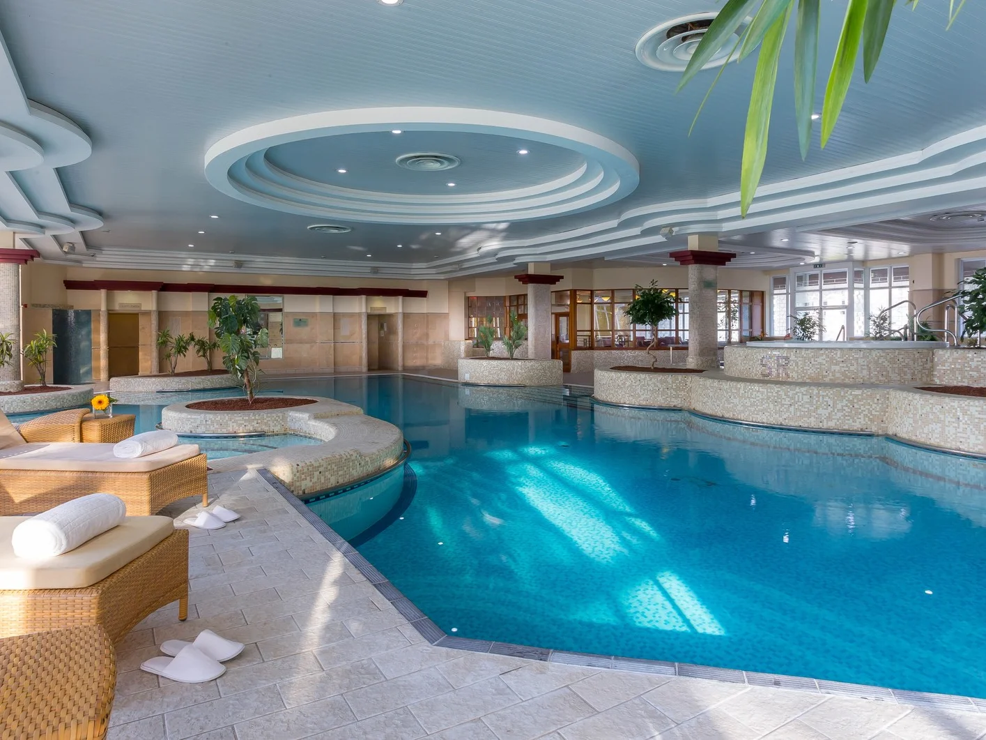 Luxury hotel indoor swimming pool with wicker sun loungers, white towels, and tropical plants