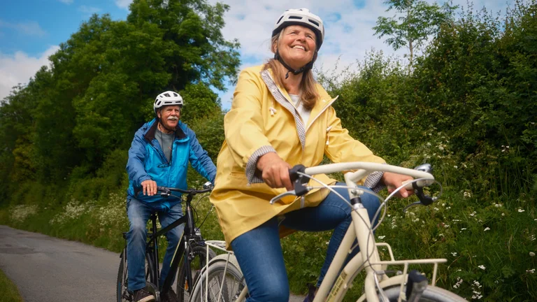 Two cyclists riding along a tree-lined greenway on a sunny day, both wearing helmets