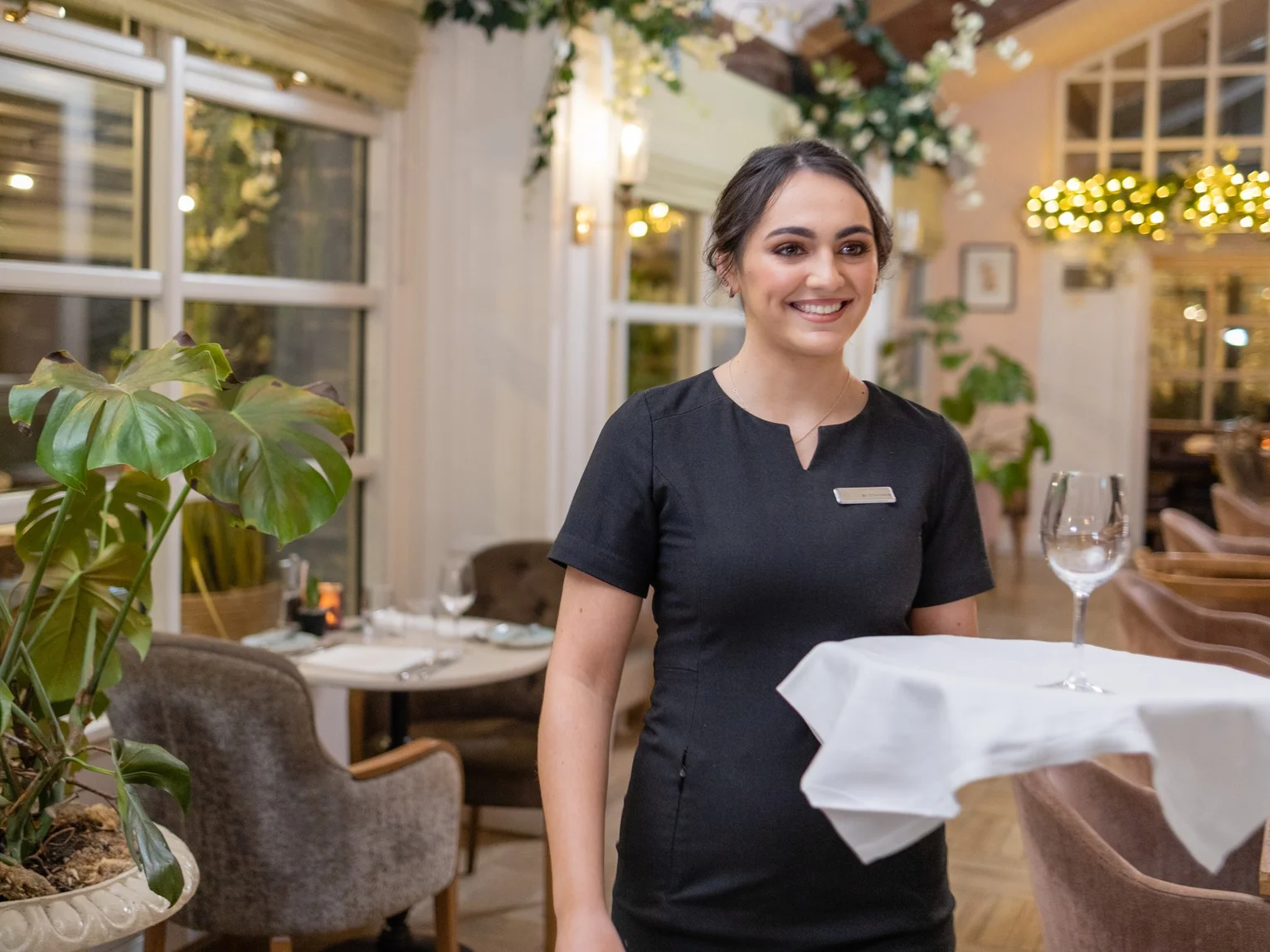 Hospitality staff member in uniform smiling while smoothing a white tablecloth on a set table in a bright restaurant