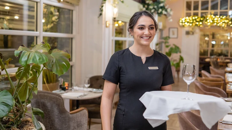 Hospitality staff member in uniform smiling while smoothing a white tablecloth on a set table in a bright restaurant
