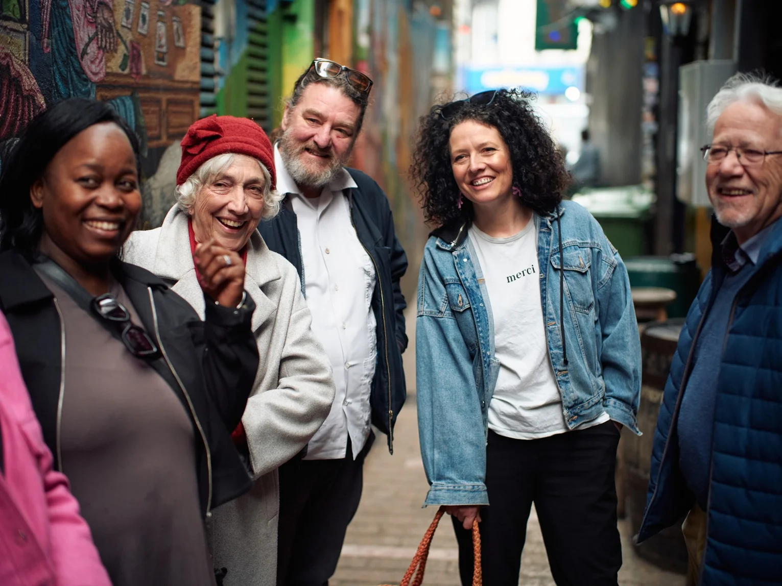 A group of five people of mixed ages laughing together in a colourful street art laneway