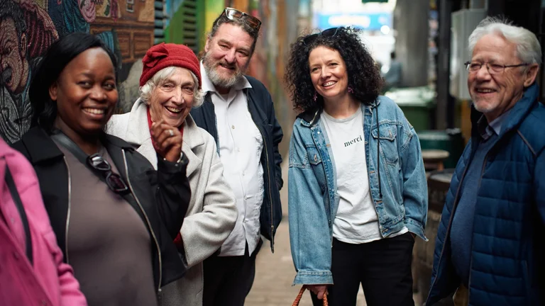 A group of five people of mixed ages laughing together in a colourful street art laneway