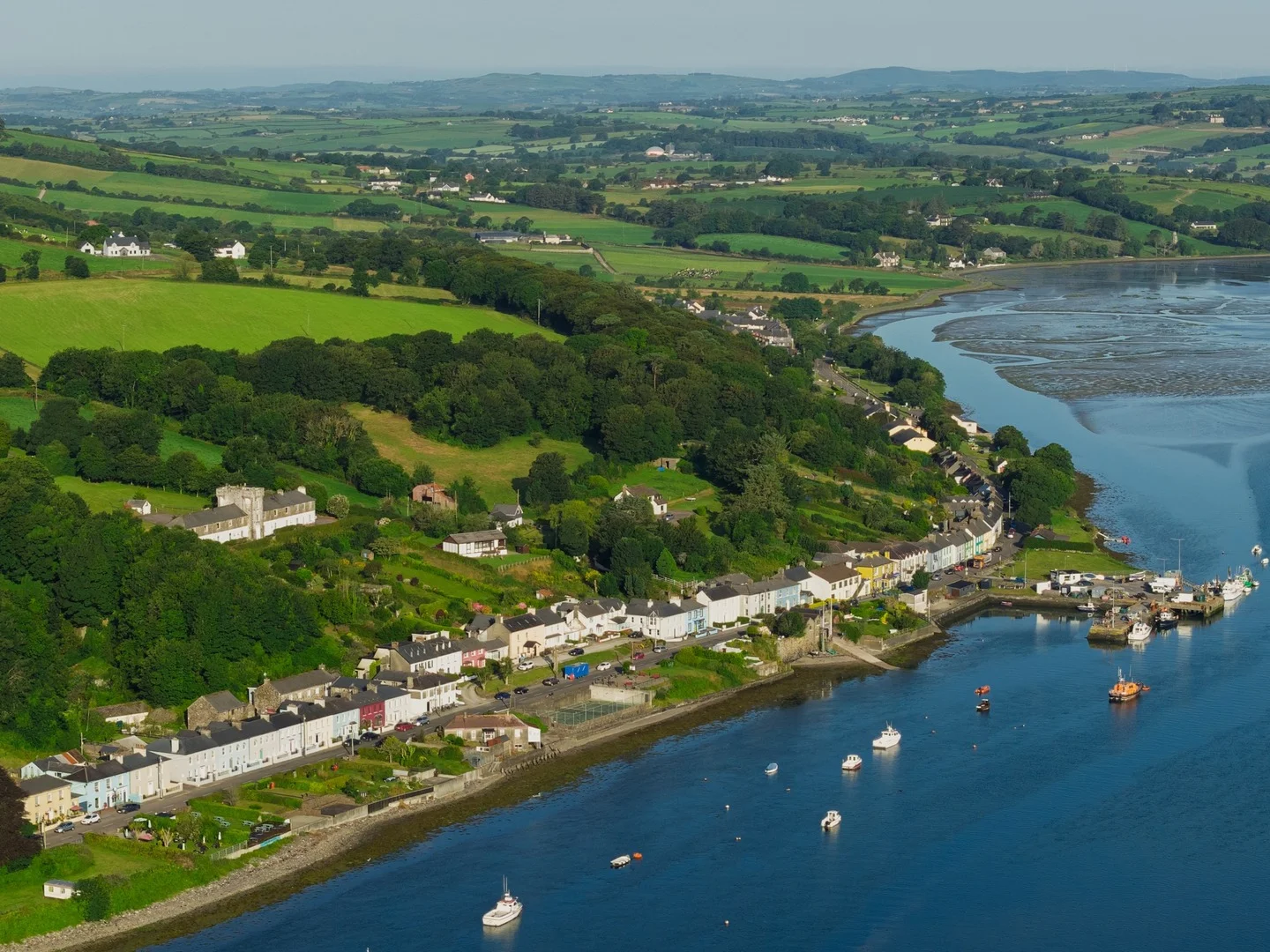 Aerial view of an Irish coastal village with colourful houses, a harbour and green countryside.