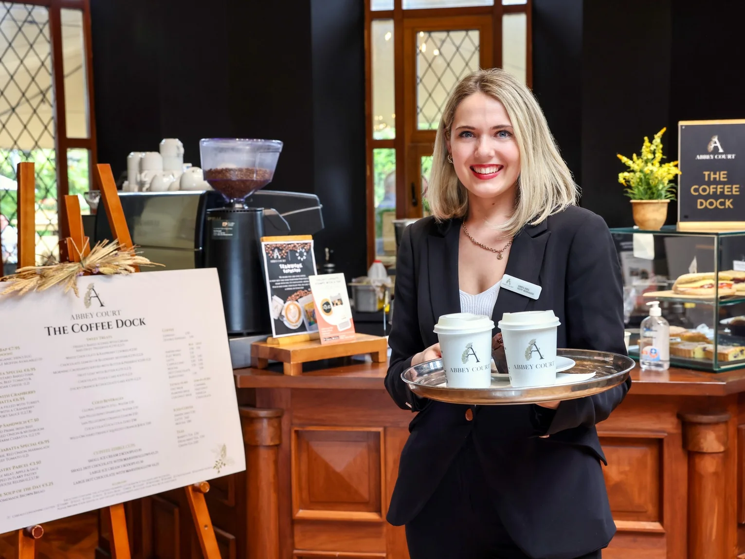 Smiling hotel staff member holding a tray of branded takeaway coffees at a café counter with menu board