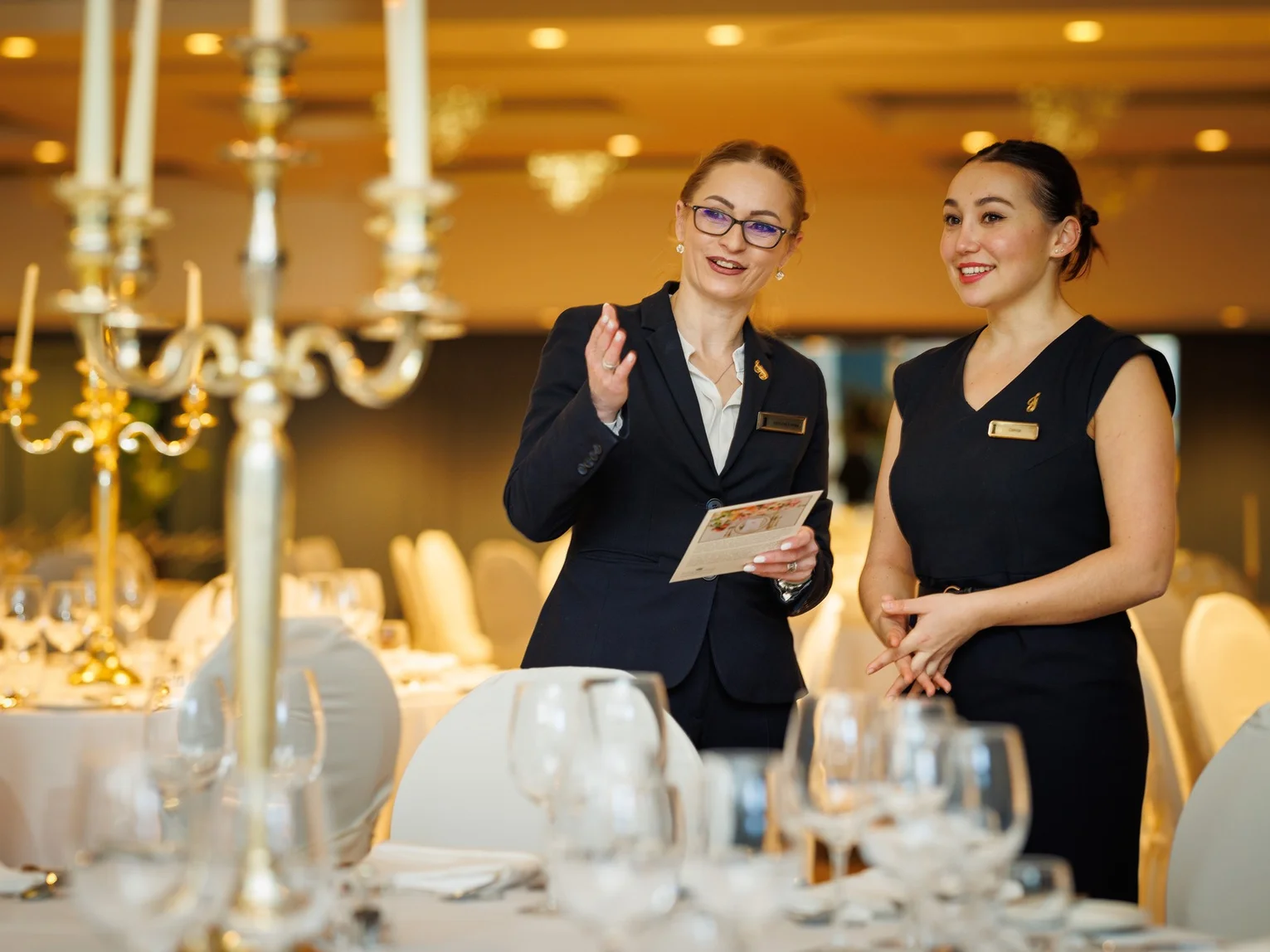 Two hotel staff members in formal uniforms speaking together in a set banquet dining room