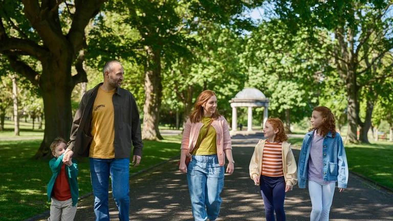 Family of five walking together along a tree-lined path in a public park on a sunny day