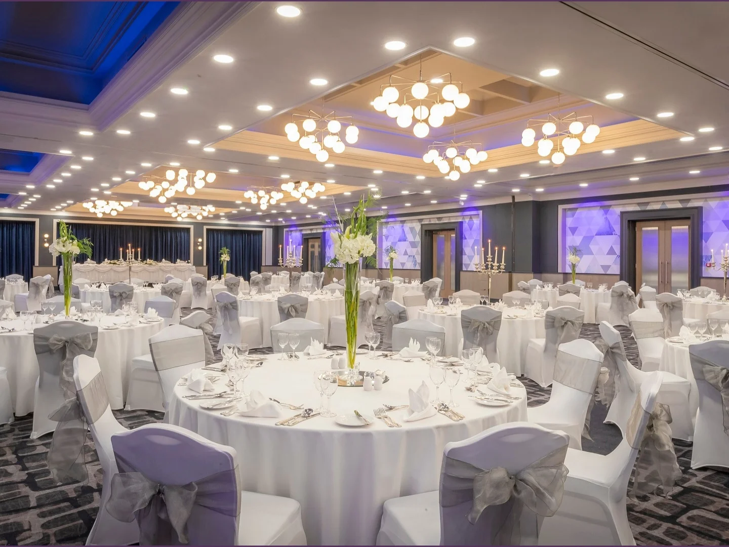 Large hotel ballroom set for a gala event, with round tables, white chair covers, grey sashes, and purple ambient lighting