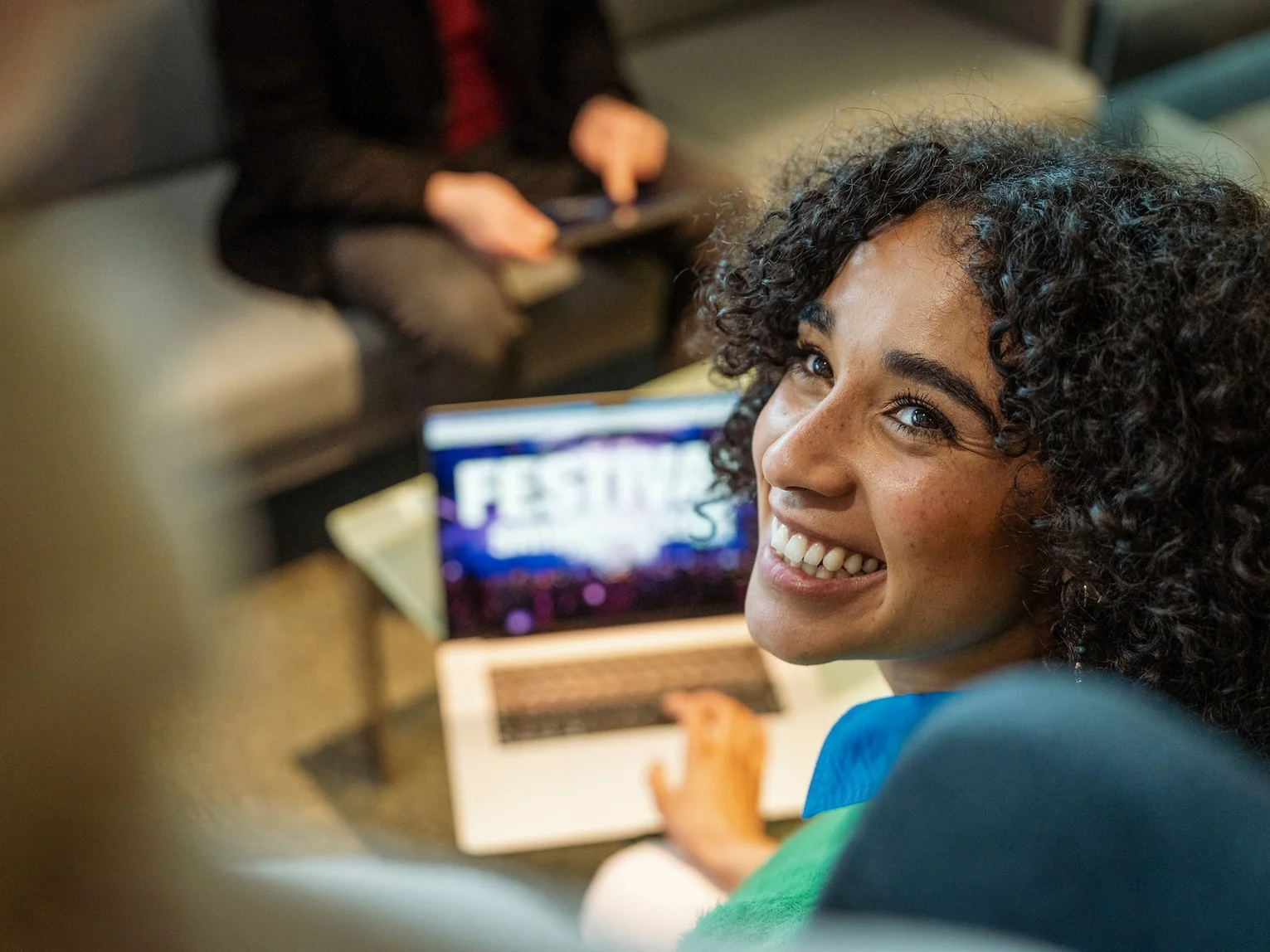 Young woman smiling while working on a laptop showing a festival website in a relaxed lounge setting