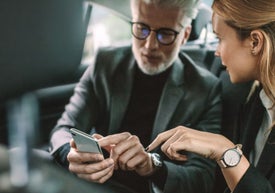 Mężczyzna w średnim wieku i kobieta siedzą w samochodzie. Mężczyzna trzyma w ręku telefon i pokazuje kobiecie coś na ekranie. Oboje są ubrani w formalne stroje.