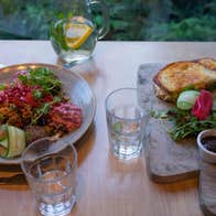Two plates full of food; one a salad with cucumber and beets, the other a toasted sandwich, all served with lemon water.