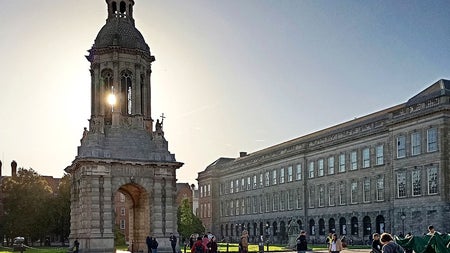 The Campanile at Trinity College Dublin with sunlight behind it and people walking across the cobblestone courtyard