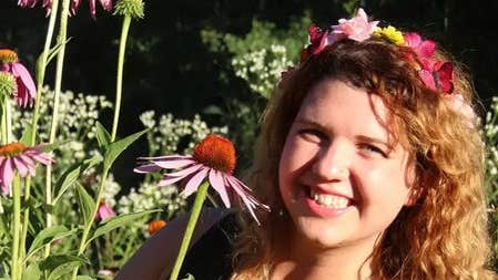 A smiling woman holding a flower, surrounded by flowering plants.