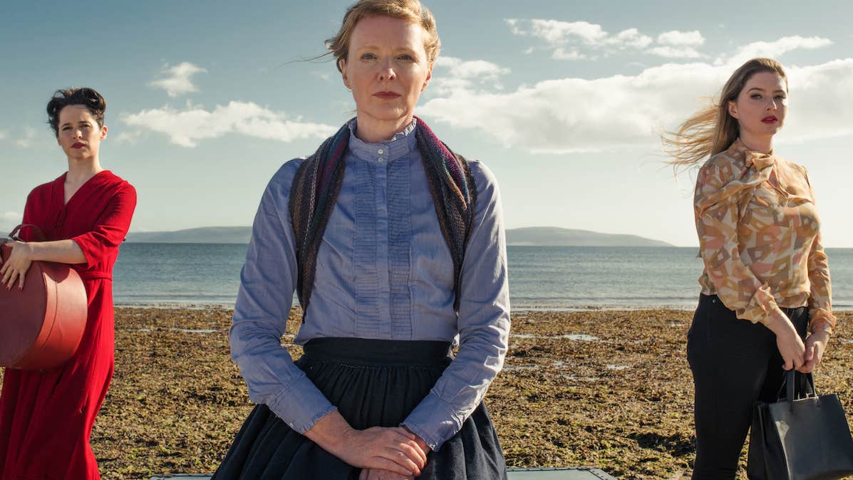 3 women standing apart on a beach with sea in background.