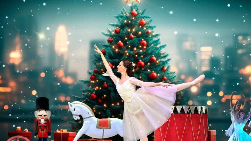 A woman in white dress is standing on one leg in ballet pose with other leg and arm outstretched, with christmas tree and toys behind.
