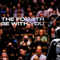 A figure in black shiny darth vadar outfit with a conductor's baton against background of an audience