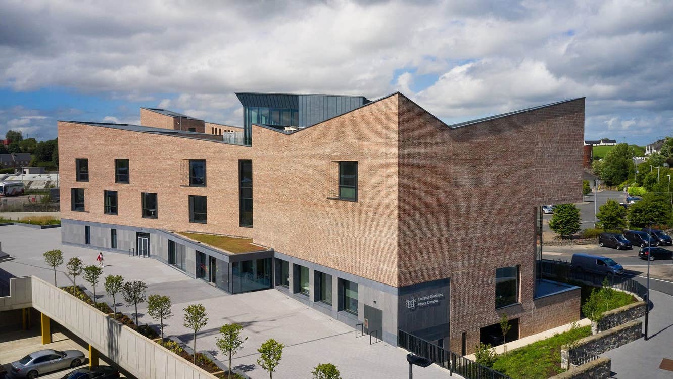 Grey and light brown building with trees by a path and cars parked outside
