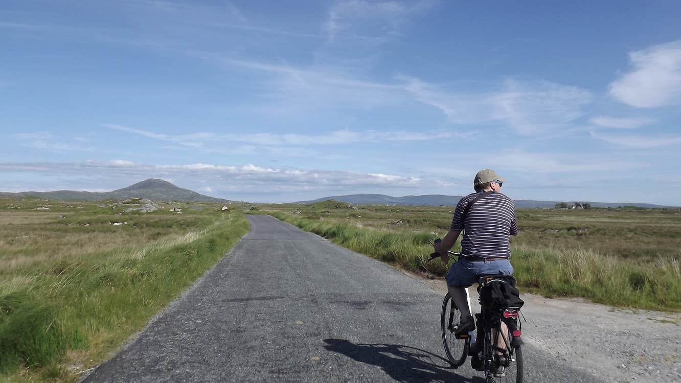 Person cycling on a road with fields either side and mountains in the background