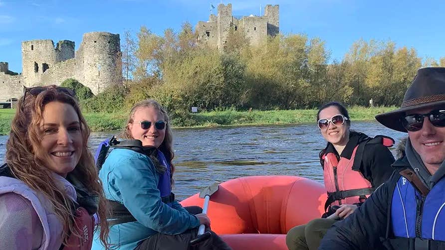 4 smiling people wearing lifejackets on red inflatable boat on water with castle in the background