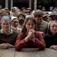 A worried woman resting her elbows on wooden stage at the front of crowd of sad looking people in period costumes.