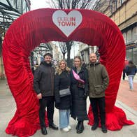 Friends pose beneath a giant red velvet heart arch in the heart of Dublin, part of the city's playful embrace of romance and love around Valentine's season, with the Gaiety Theatre visible in the background.