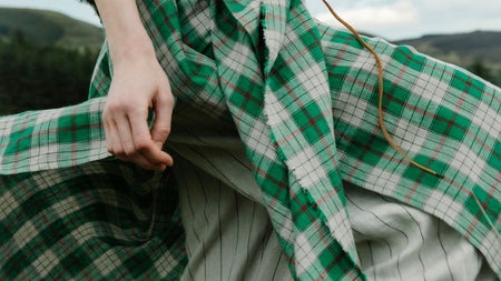 A close up of a lady wearing a large green tartan wrap around type of clothing standing on grass outdoors