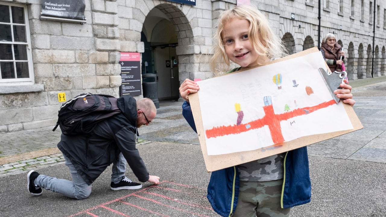 Outside an old building with many arches, a young child is proudly holding a drawing with a man on his knee behind, drawing with chalk on the concrete ground.