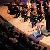 A smiling man in a colourful shirt is seated on the edge of a stage waving at the audience with an orchestra playing behind him.