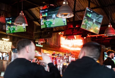 Two people in a pub with a rugby match showing on three screen above the bar
