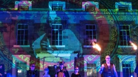 Samhain Chiarraí Street Spectacle, a line of people at night in costumes standing in front of a building lit up with logo from light show.
