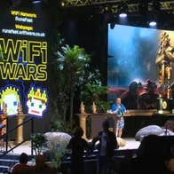 WiFi Wars: Interactive Comedy Game Show, view of a stage with a man talking, a large screen to one side.