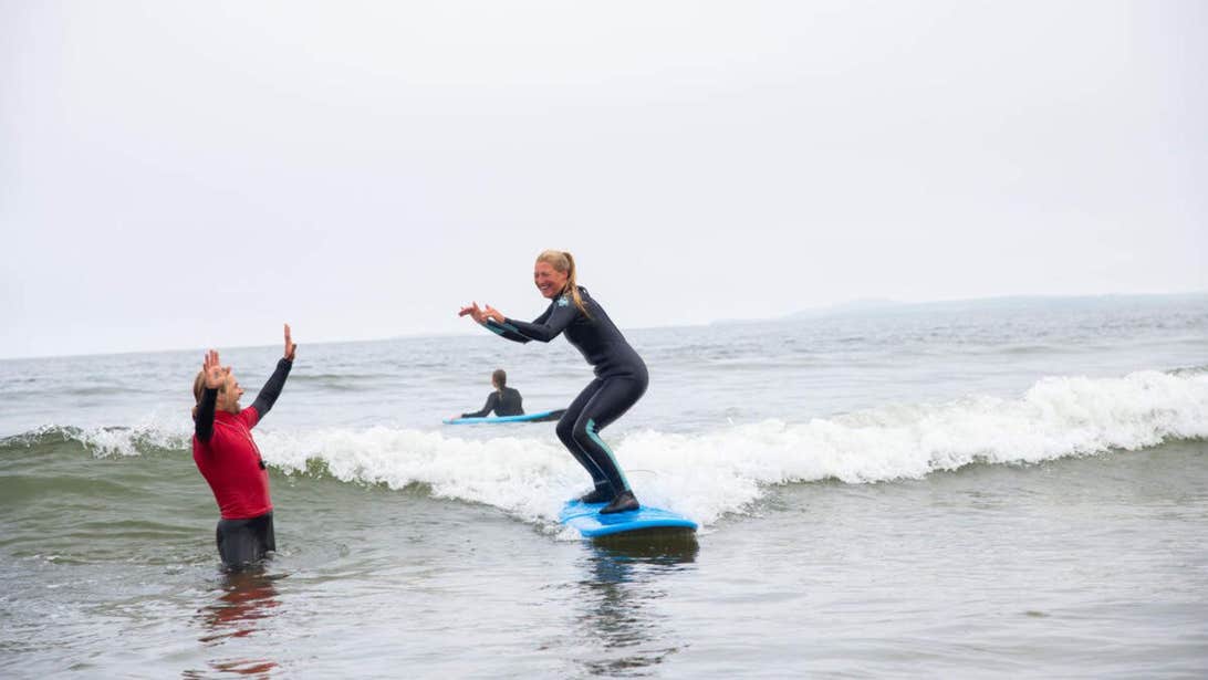 Woman learning to surf on surf board while the male teacher stands in the water cheering her on.