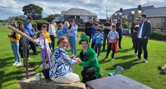 Lady sitting on a rock with a man in a leprechaun costume kneeling down and people in the background