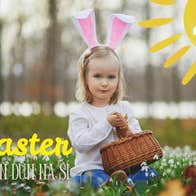 A young girl with rabbit ears headband holding a wicker basket seated on the ground with small white flowers.