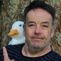 Púca – John Colleary: Mighty Quack, a close up of a man pointing over his shoulder at a white toy duck behind him