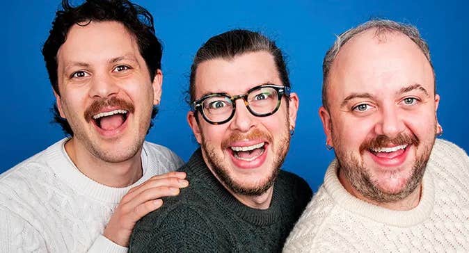 3 laughing men in a tight line wearing aran style knitted jumpers against plain blue background.