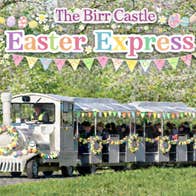 The Birr Castle Easter Express - a white train on a road pulling 2 decorated carriages in eggs and flowers in a woodland setting with blossom.