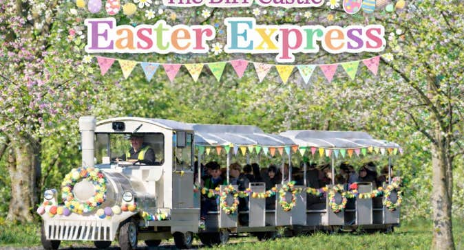 The Birr Castle Easter Express - a white train on a road pulling 2 decorated carriages in eggs and flowers in a woodland setting with blossom.