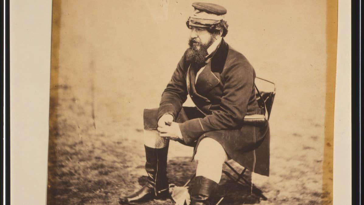 An old, brown style photo of a man in uniform with black boots seated on a camping chair.