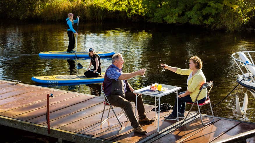 A couple having a meal beside a waterway in Leitrim with two people on stand-up paddleboards in the background