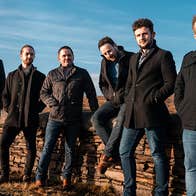 A group of 6 men in coats are standing in a line on the edge of a road with bogland behind.