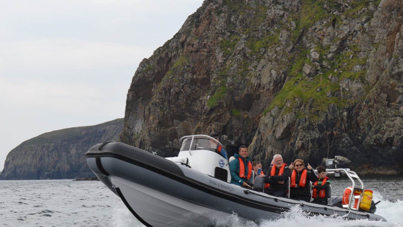 People on a grey rib boat in the water wearing orange life jackets in front of cliffs