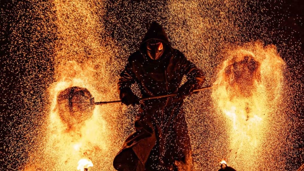 A dark hooded figure against background of flames and sparks from burning.