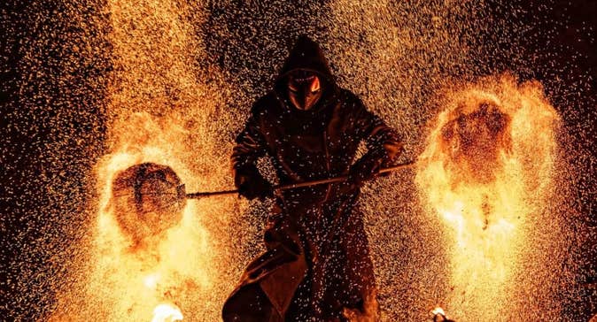 A dark hooded figure against background of flames and sparks from burning.
