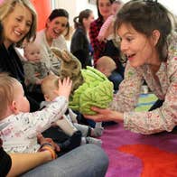 A woman is smiling and holding up a soft toy of a rabbit in a lettuce to a small baby on a parent's lap with others looking on.