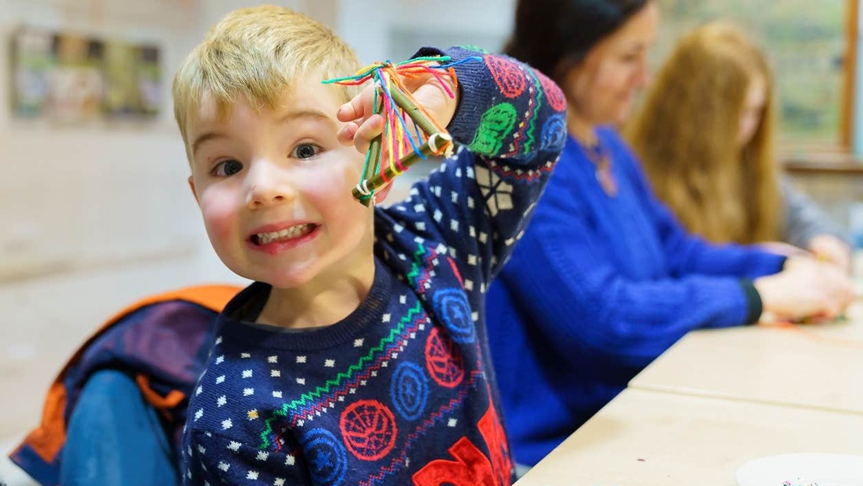A smiling young boy in a Christmas jumper seated at a table holding up a small handmade decoration